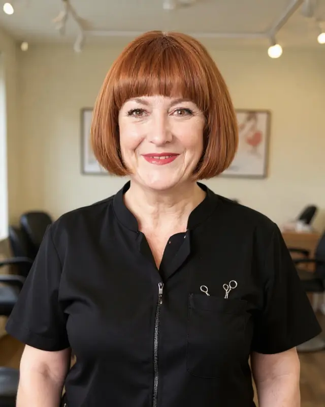 Jackie Holroyd, an AI character: a woman with a copper-red bob in a black zip-front tunic, scissors in her pocket, standing in her salon.